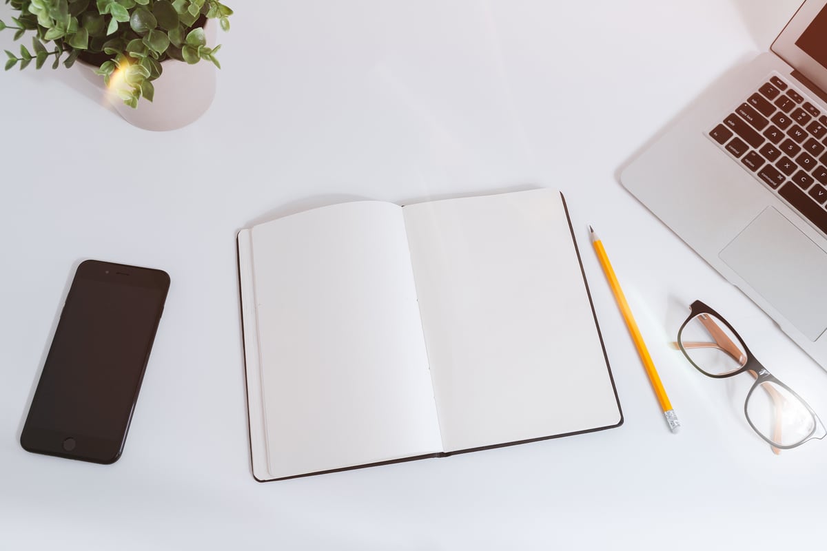 A smartphone beside an open notebook, a pencil, glasses and a laptop on a white desk