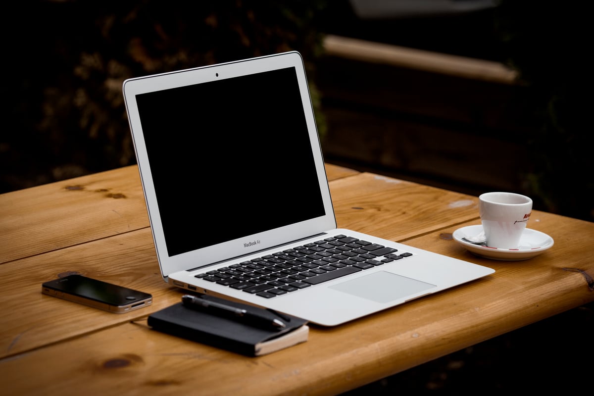 An open laptop, a phone, a notebook and a coffee cup on a wooden table
