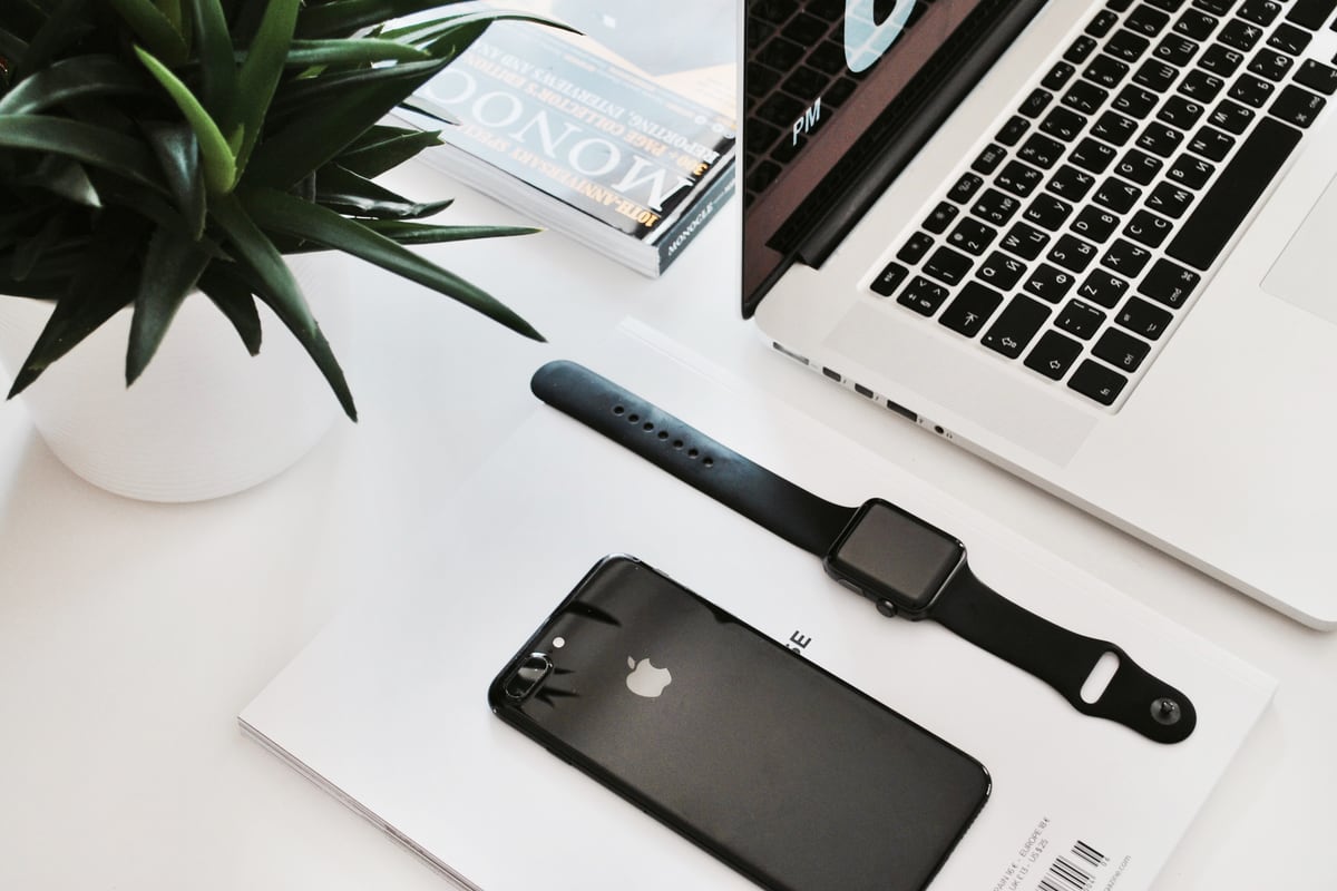 A smartphone and a smartwatch on a white desk beside a laptop and a potted plant